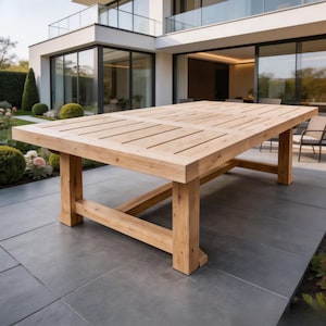 May include: A large, rectangular wooden outdoor dining table. The table has a light brown finish and a slatted tabletop design. The legs are thick and sturdy, providing a solid base. The table is set on a gray patio, with a modern house in the background.
