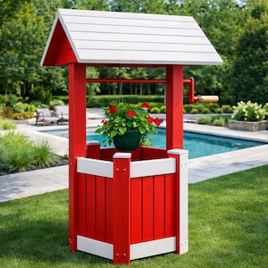 May include: A red and white decorative wishing well with a white roof and a hanging green flower pot filled with red flowers. The well has a wooden handle and is set on a green lawn near a swimming pool.