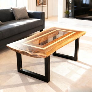 May include: A rectangular wooden coffee table with a glass insert and a natural edge. The table has a light brown finish with a gold-colored border around the glass. It is supported by black metal legs.