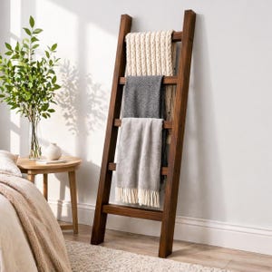 May include: A dark brown wooden blanket ladder with three blankets draped over the rungs. The blankets are cream, grey, and light grey with fringe. The ladder is leaning against a white wall.