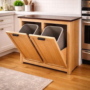 Könnte beinhalten: Ein Küchenschrank aus Holz mit zwei Fächern, in denen sich jeweils ein grauer Mülleimer befindet. Die Schranktüren sind geöffnet und geben die Behälter frei. Der Schrank ist aus hellem Holz mit einer dunkelbraunen Arbeitsplatte.