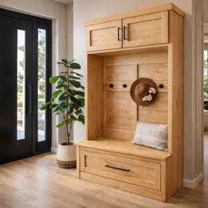 May include: A light oak wood entryway bench with a drawer, hooks, and upper storage cabinets. A brown hat with floral accents hangs on the wall. A decorative pillow rests on the bench. A potted plant and black door are in the background.