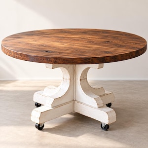 Op de afbeelding: Een ronde eettafel met een donkerbruin gebeitst houten blad met zichtbare nerven en noesten. Het onderstel is gemaakt van wit geschilderd hout met een verweerde afwerking, ondersteund door vier stevige poten, elk voorzien van een zwart zwenkwiel.