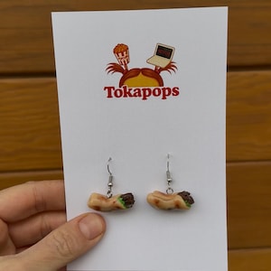 Könnte beinhalten: Handgefertigte Ohrringe in Form von Mini-Burritos. Die Ohrringe sind an silberfarbenen Fischhaken befestigt. Sie werden auf einer weißen Karte mit dem Text "Tokapops" und einem Logo mit Popcorn und einem Laptop präsentiert.