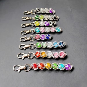May include: A collection of eight keychains featuring silver-toned metal lobster clasps attached to chains of interlocking silver rings. Each chain holds a series of round, multi-colored beads in various sequences, including solid and translucent colors.