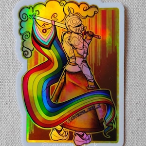 May include: A sticker featuring a knight in golden armor holding a sword with a rainbow flag. The knight is set against a gold background with a rainbow ribbon flowing from the flag. The sticker has the text "@reverie_art_k".