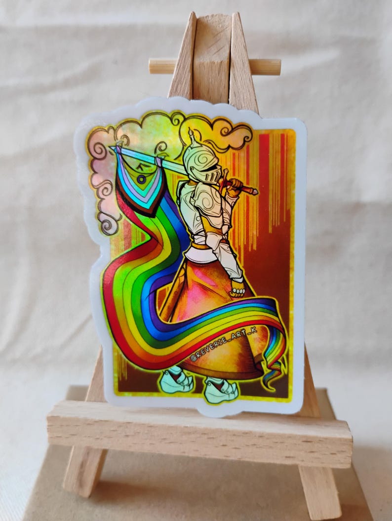 May include: A holographic sticker featuring a knight holding a rainbow flag and sword. The knight is wearing armor and a helmet. The sticker has a white border with a cloud design. The background is a gradient of gold and brown.