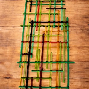 May include: Abstract wall art piece with a grid-like design. The artwork features a mix of colorful glass rods in green, yellow, red, and black, arranged in a layered pattern. A silver chain is attached for hanging.