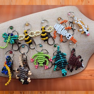 May include: A collection of beaded animal keychains, including a dragonfly, bee, frog, lizard, and turtle. The keychains are made with various colors of beads and feature silver-tone key rings and clasps. The keychains are displayed on a beige fabric.