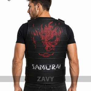 Może przedstawiać: Czarna kamizelka z poziomymi wyściełanymi segmentami i czerwoną grafiką stylizowanej twarzy na plecach. Słowo SAMURAI jest nadrukowane białymi, postarzanymi literami pod grafiką. Kamizelka jest założona na czarny t-shirt z krótkim rękawem.
