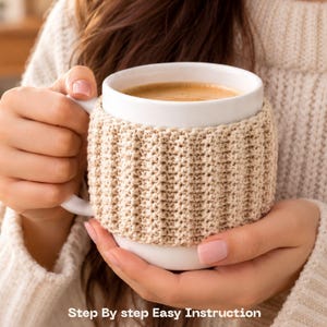 Può includere: Una tazza in ceramica bianca con un rivestimento all'uncinetto beige a coste. La tazza è riempita con una bevanda al caffè marrone chiaro. Il testo "Step By step Easy Instruction" è stampato in bianco nella parte inferiore dell'immagine.