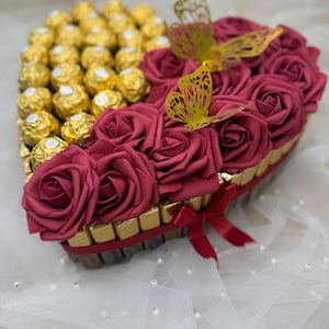 Könnte beinhalten: Herzförmiges Arrangement mit goldverpackten Pralinen auf der einen und burgunderroten Rosen auf der anderen Seite. Goldene Schmetterlingsausschnitte und ein rotes Band sorgen für dekorative Akzente. Das Arrangement liegt auf einer weißen Oberfläche.