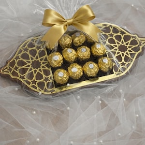 Könnte beinhalten: Eine dekorative goldfarbene Schale gefüllt mit Schokoladenbonbons, die in Goldfolie verpackt sind. Die Schale ist in durchsichtiges Cellophan gewickelt und mit einem goldenen Band gebunden. Die Schale hat ein geometrisches Design.