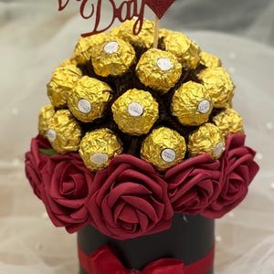 Könnte beinhalten: Valentinstagsgeschenk-Arrangement. Eine schwarze Box mit rotem Band und Rosen. Die Anordnung ist mit goldverpackten Pralinen und einem glitzernden "Happy Valentine's Day"-Schild versehen.
