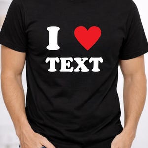 Könnte beinhalten: Schwarzes T-Shirt mit dem Text "I ♥ TEXT" in Weiß und einem roten Herz. Das Shirt hat einen Rundhalsausschnitt und wird von einer Person getragen. Die Person trägt eine blaue Jeans und hat die Hände in den Taschen.