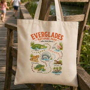 May include: A beige canvas tote bag featuring a colourful graphic print of Everglades National Park wildlife. The design includes illustrations of an alligator, manatee, sea turtle, wading bird, and otter, with the text Everglades National Park.