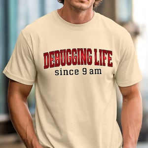 Könnte beinhalten: Beiges T-Shirt mit dem Text "DEBUGGING LIFE" in roter Schrift mit schwarzer Umrandung und "since 9 am" in Schwarz. Das Shirt hat kurze Ärmel und einen Rundhalsausschnitt.
