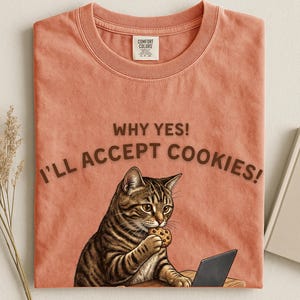 Könnte beinhalten: Korallenfarbenes T-Shirt mit einer Grafik einer getigerten Katze, die einen Keks isst, während sie einen Laptop benutzt. Der Text über der Katze lautet: "WHY YES! I'LL ACCEPT COOKIES!" Das Shirt hat ein Comfort Colors-Etikett.