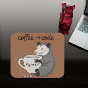 Op de afbeelding: Een bruine muismat met de tekst "coffee -> code" en een cartoon opossum die een koffiekopje vasthoudt met "espresso" erop. Een rode gummybeer en een laptop zijn zichtbaar.