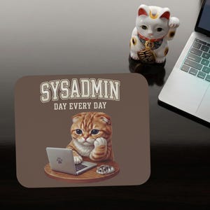 Op de afbeelding: Bruine muismat met de tekst "SYSADMIN DAY EVERY DAY" en een cartoonkat die een laptop gebruikt. Een wit gelukskatbeeldje en een laptop zijn ook zichtbaar.