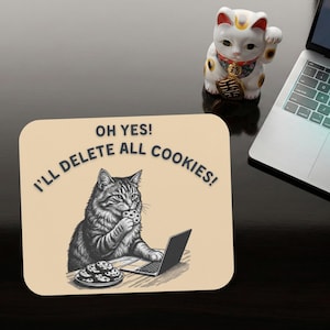 Pode incluir: Um tapete de rato bege com uma ilustração de um gato comendo um biscoito enquanto usa um laptop. O texto no tapete do rato diz "OH YES! I'LL DELETE ALL COOKIES!". Uma estatueta de gato da sorte branca e um laptop também são visíveis.