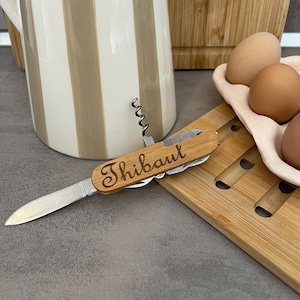 Peut inclure: Un couteau suisse multifonction avec un manche en bois clair portant le nom gravé Thibaut. L'outil comprend une lame en métal, un tire-bouchon et d'autres ustensiles pliables, présentant un design fonctionnel pour diverses tâches en cuisine.