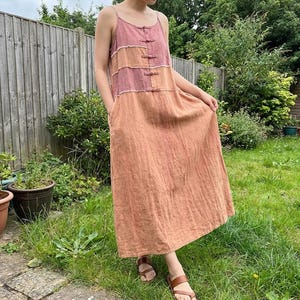 Puede incluir: Un vestido largo sin mangas en tonos rosa y naranja. El vestido presenta un diseño escalonado con cierres de botones y tirantes finos. La modelo lleva sandalias marrones. El vestido está hecho de una tela texturizada.