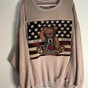 May include: A tan sweatshirt with a tapestry-like design featuring a teddy bear saluting in front of an American flag. The words "GOD BLESS AMERICA" are displayed. The sweatshirt has ribbed detailing on the sides.