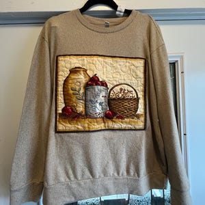 May include: A beige long-sleeve sweatshirt featuring a rectangular quilted panel on the front. The panel displays a still life illustration of a tall tan jug, a blue canister with red apples, and a woven basket filled with berries.