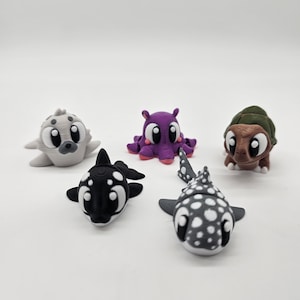 Puede incluir: Colección de cinco figuras caprichosas de criaturas marinas impresas en 3D. El conjunto incluye una foca gris, un pulpo morado, una orca en blanco y negro, un pez gris y blanco moteado y una tortuga marrón. Cada figura tiene ojos grandes y de dibujos animados.