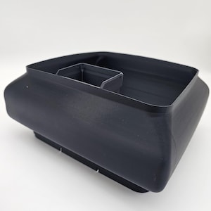 May include: A black, rectangular container with a built-in divider. The container has a slightly curved design and sits on a base with small cutouts. The overall shape is boxy with rounded corners.