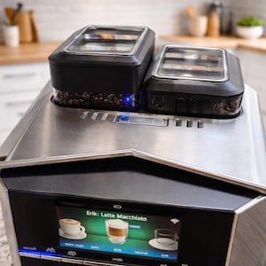 May include: Stainless steel espresso machine with two black bean containers on top. The machine has a digital display showing a latte macchiato and other options. The brand name "SIEMENS" is visible.