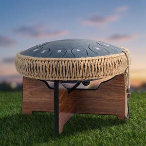 Pode incluir: Um tambor de língua de aço com um topo azul-acinzentado e uma borda de corda natural, colocado em um suporte de madeira. O tambor tem várias notas recortadas e é projetado para performance musical. O suporte é feito de madeira escura.