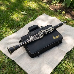 May include: A black clarinet with silver keys rests on top of a black case. The case is on a beige cloth, set on a bed of green grass. The clarinet is angled, showcasing its keys and bell. The case has the text "LADE Designed in USA".