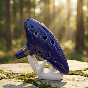 May include: A deep blue ocarina with multiple finger holes, resting on a white ceramic hand-shaped stand. The musical instrument has a glossy finish and a silver-colored band near the mouthpiece. The background is a blurred view of a forest.