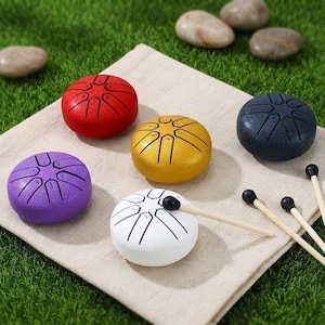 May include: A collection of colorful steel tongue drums in red, gold, navy blue, purple, and white. Each drum has etched lines and numbers. Wooden drumsticks with black tips are also present, resting on a beige cloth.