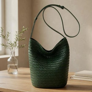 Peut inclure: Un sac à bandoulière en cuir tressé vert foncé avec une longue sangle. Le sac est cylindrique avec une large ouverture. Il est posé sur une table en bois près d'une fenêtre et d'un vase avec de la verdure.