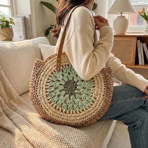 May include: A circular woven straw tote bag with a brown leather handle. The bag has a circular pattern in shades of green, brown, and beige. The bag is designed for carrying items and has a casual, bohemian style.
