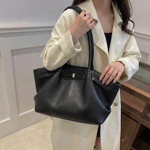Può includere: Borsa tote in pelle nera con finitura strutturata. La borsa ha due manici e una chiusura color oro. La borsa è tenuta da una persona che indossa un cappotto color crema.