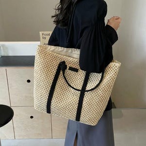 May include: A woven beige tote bag with black handles and accents. The bag has a rectangular shape and a spacious interior. The handles are thick and knotted at the top. The bag is being held by a person wearing a black top and a gray skirt.