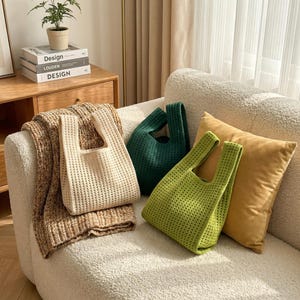 May include: Three knitted tote bags in cream, dark green, and lime green, displayed on a white sofa with a mustard-yellow cushion. A brown knitted throw and a small potted plant are also visible. The bags have an open weave design.