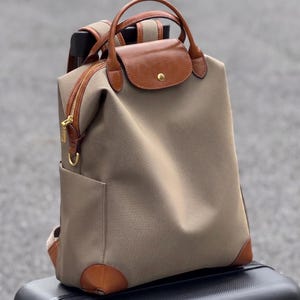 May include: A taupe-colored backpack featuring brown leather trim, a top handle, and a fold-over flap with a gold-tone snap closure. The bag includes a gold-tone zipper along the side, adjustable shoulder straps, and reinforced leather corners.