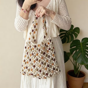 Könnte beinhalten: Eine beige Makramee-Tragetasche mit Gittermuster, mit braunen, orangefarbenen und beigen Perlen. Die Tasche hat zwei Henkel und wird von einer Person in einem weißen Kleid gehalten. Eine Topfpflanze befindet sich im Hintergrund.