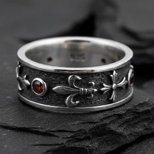 Op de afbeelding: Een zilveren ring met een donkere, getextureerde band met fleur-de-lis- en kruisontwerpen. Kleine, ronde, rode edelstenen zijn in de band gezet. De ring wordt gepresenteerd op een donkere, getextureerde ondergrond.
