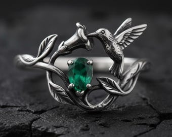 May include: A silver ring featuring a hummingbird design. The ring has a pear-shaped, emerald-green gemstone at the center, surrounded by a floral and leaf motif. The hummingbird is depicted in flight, with its beak near a flower.