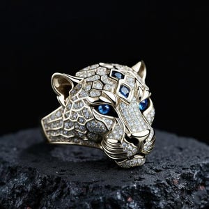 May include: A gold-coloured ring in the shape of a leopard's head, adorned with tiny diamonds and blue gemstones for the eyes. The ring is displayed against a dark backdrop, emphasising its intricate details and luxurious design.