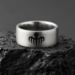 Peut inclure: Une bague argentée avec une finition brossée. La bague présente un motif graphique noir sur le devant. Elle est posée sur une surface sombre et texturée. La bague est un bijou.