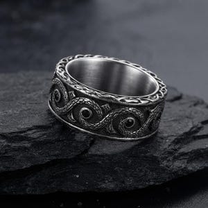 Puede incluir: Un anillo ancho de metal plateado con un patrón detallado en relieve de espirales entrelazadas similares a serpientes. El diseño incluye áreas oscuras y hundidas que resaltan la textura intrincada de las escamas y el acabado metálico general.