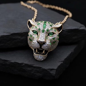 Puede incluir: Un collar dorado con un colgante de jaguar detallado. La cabeza del jaguar está adornada con brillantes piedras preciosas blancas y verdes, con gemas verdes acentuando los ojos y las manchas. El collar se presenta sobre una superficie oscura y texturizada.