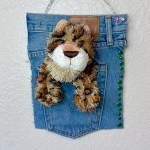 May include: A decorative wall hanging featuring a plush leopard toy peeking out of a denim pocket. The denim is a light blue wash, with a brown leather patch and green embellishments. A beaded string is used for hanging.